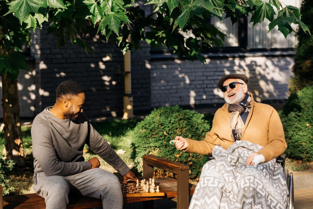 senior disabled man in wheelchair and african american cuidador playing chess together on street